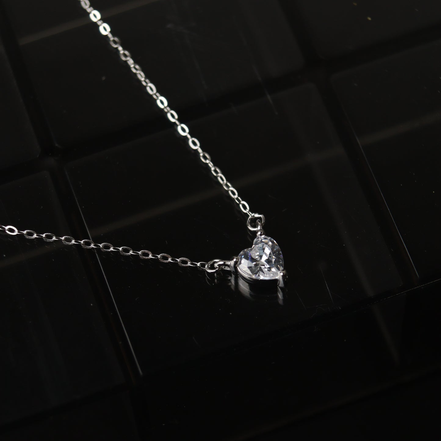 A silver necklace featuring a heart-shaped pendant, displayed on a reflective surface.