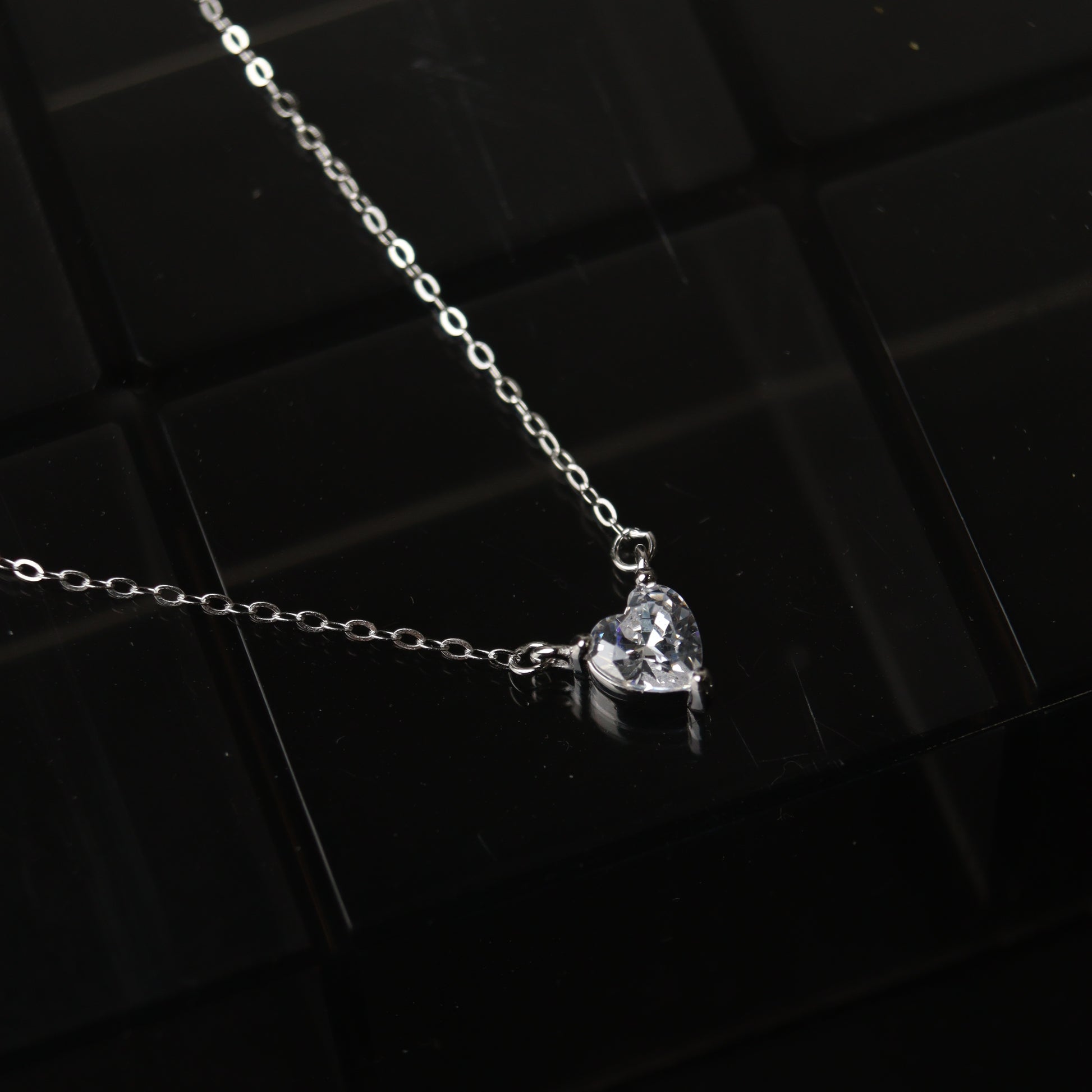 A silver necklace featuring a heart-shaped pendant, displayed on a reflective surface.